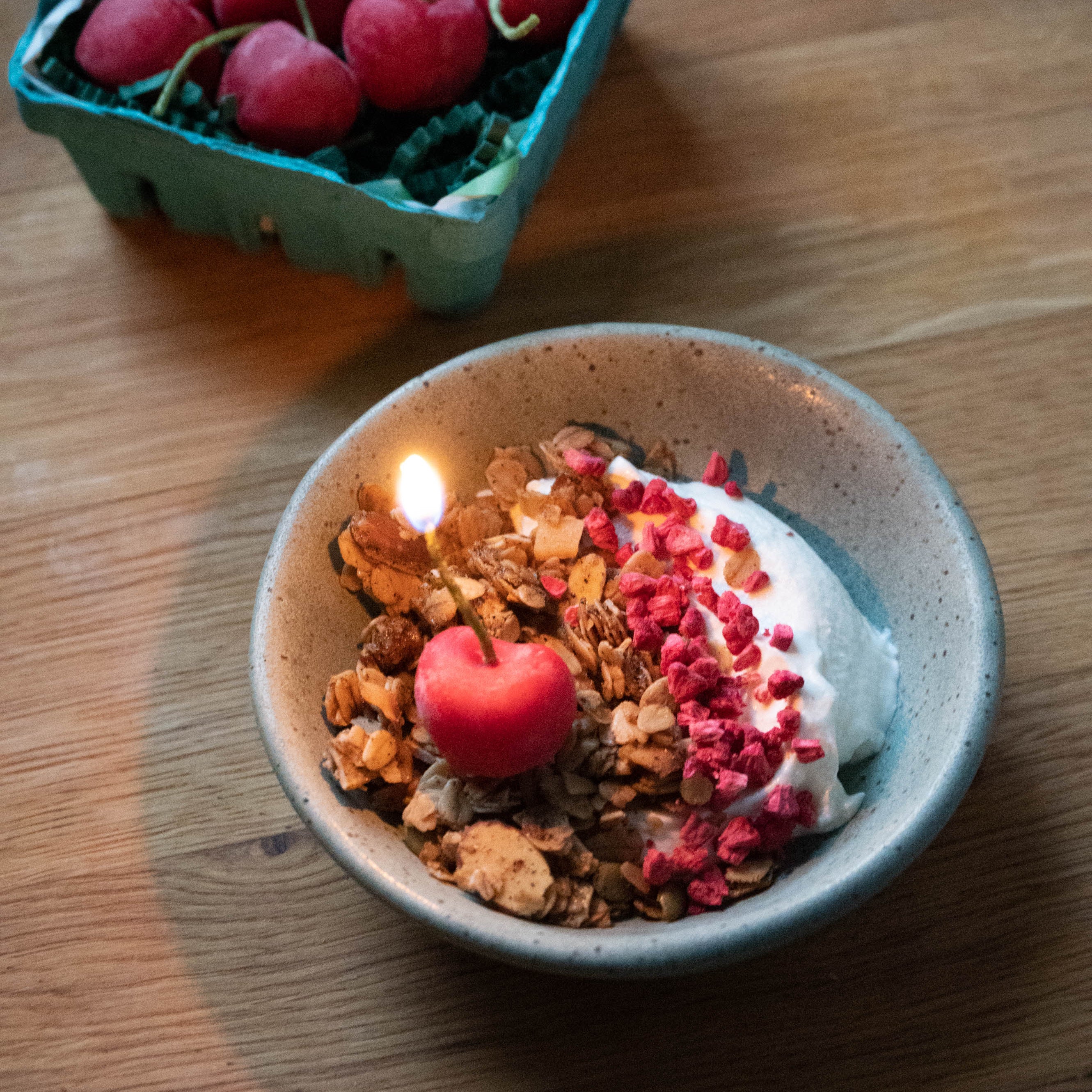 A styled food scene on a wooden table showing a bowl of granola with yogurt, dried berries, and a small red heart-shaped decoration, alongside a green container of fresh red cherries.