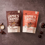 Two bags of Jamie's Farm New York Small Batch Granola are displayed on a textured gray surface, styled with chocolate pieces, sea salt, and cherries. The left bag is Dark Chocolate with Sea Salt (brown), and the right bag is Vanilla Bean with Sour Cherries (pink).