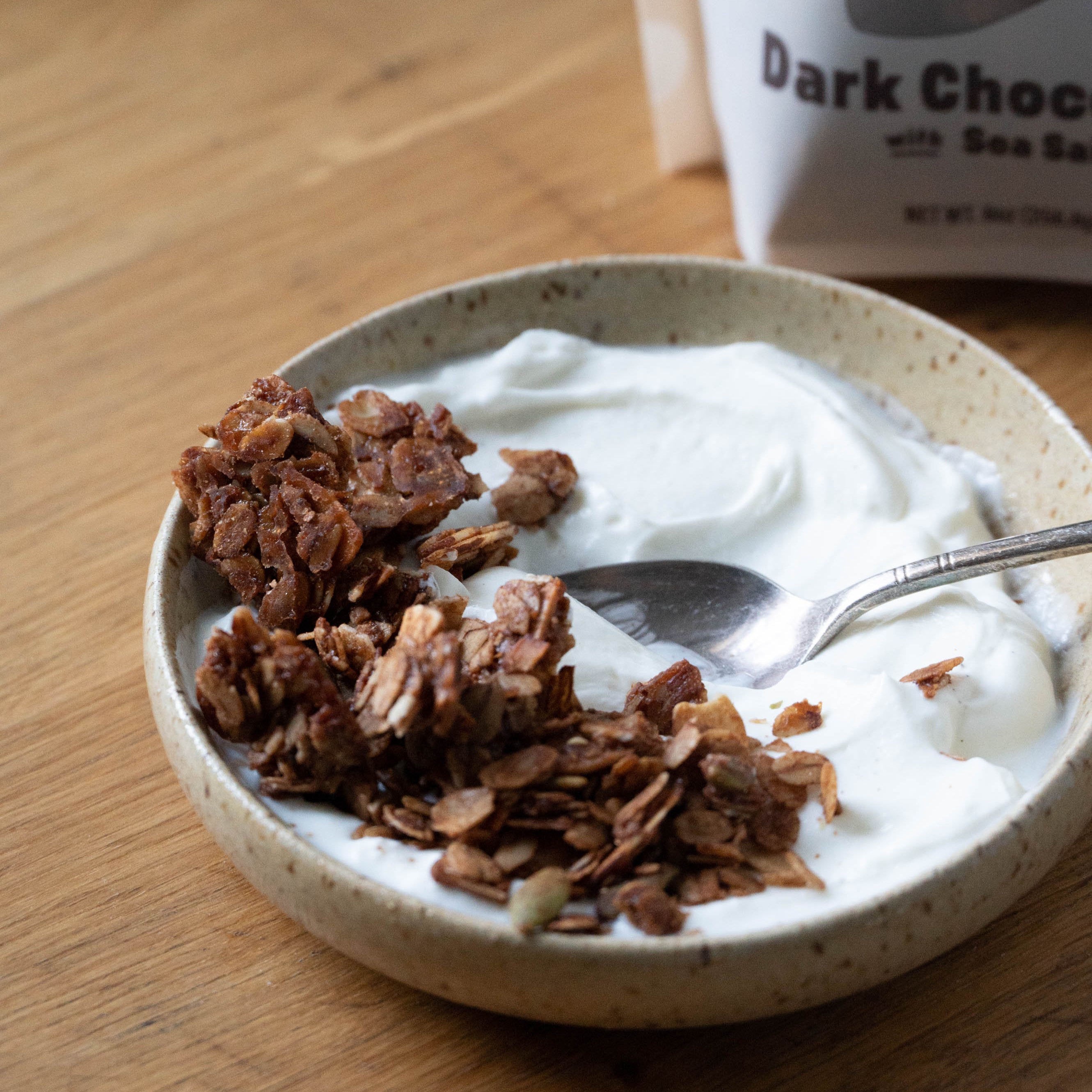 Yogurt with granola in a bowl on a wooden surface, with a package of dark chocolate in the background.