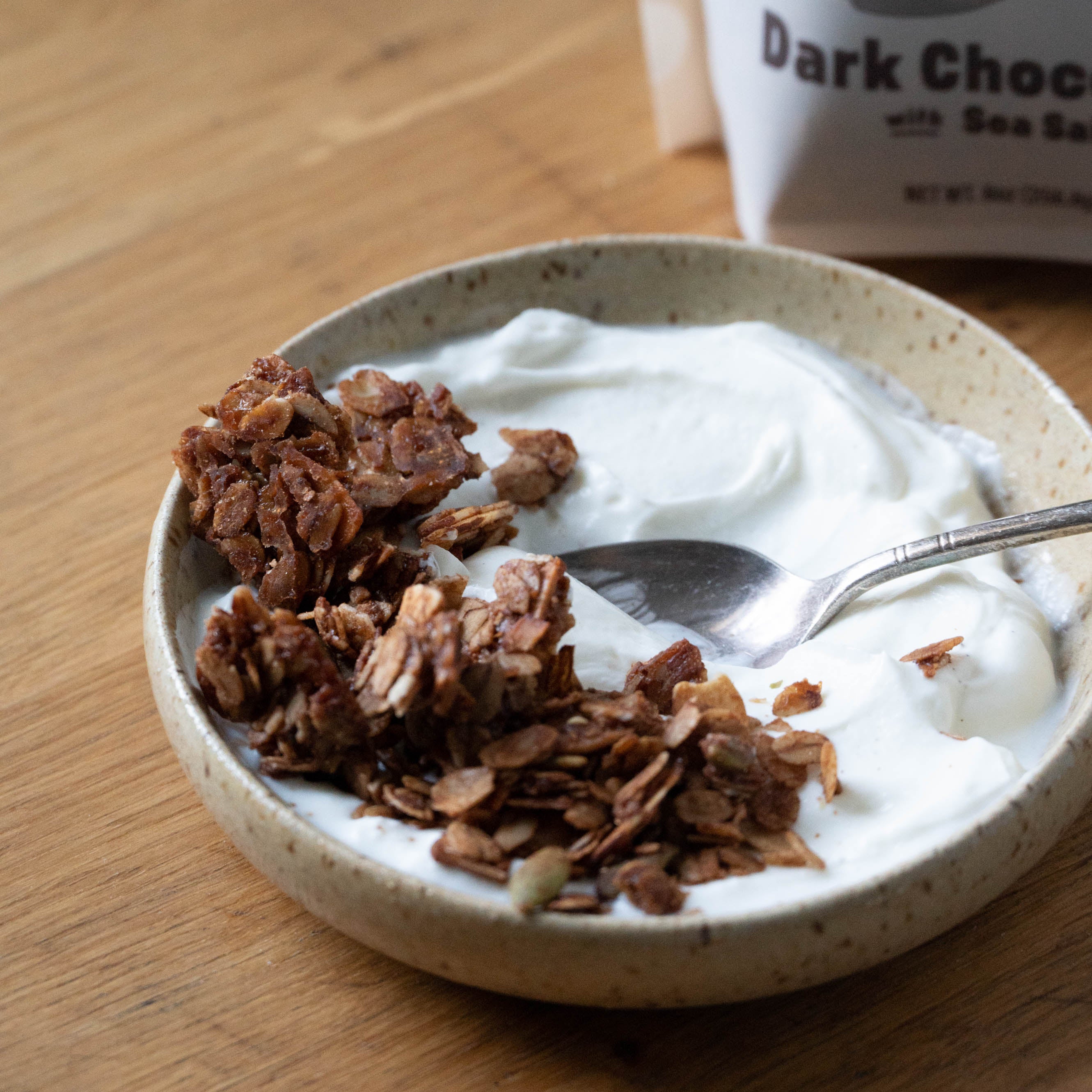 Yogurt with granola in a bowl on a wooden surface, with a package of dark chocolate in the background.