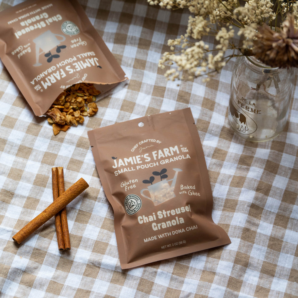 Two bags of Jamie's Farm Small Pouch Granola, specifically Chai Streusel Granola, displayed on a brown and white checkered tablecloth with cinnamon sticks and a glass jar of dried flowers.