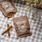 Two bags of Jamie's Farm Small Pouch Granola, specifically Chai Streusel Granola, displayed on a brown and white checkered tablecloth with cinnamon sticks and a glass jar of dried flowers.