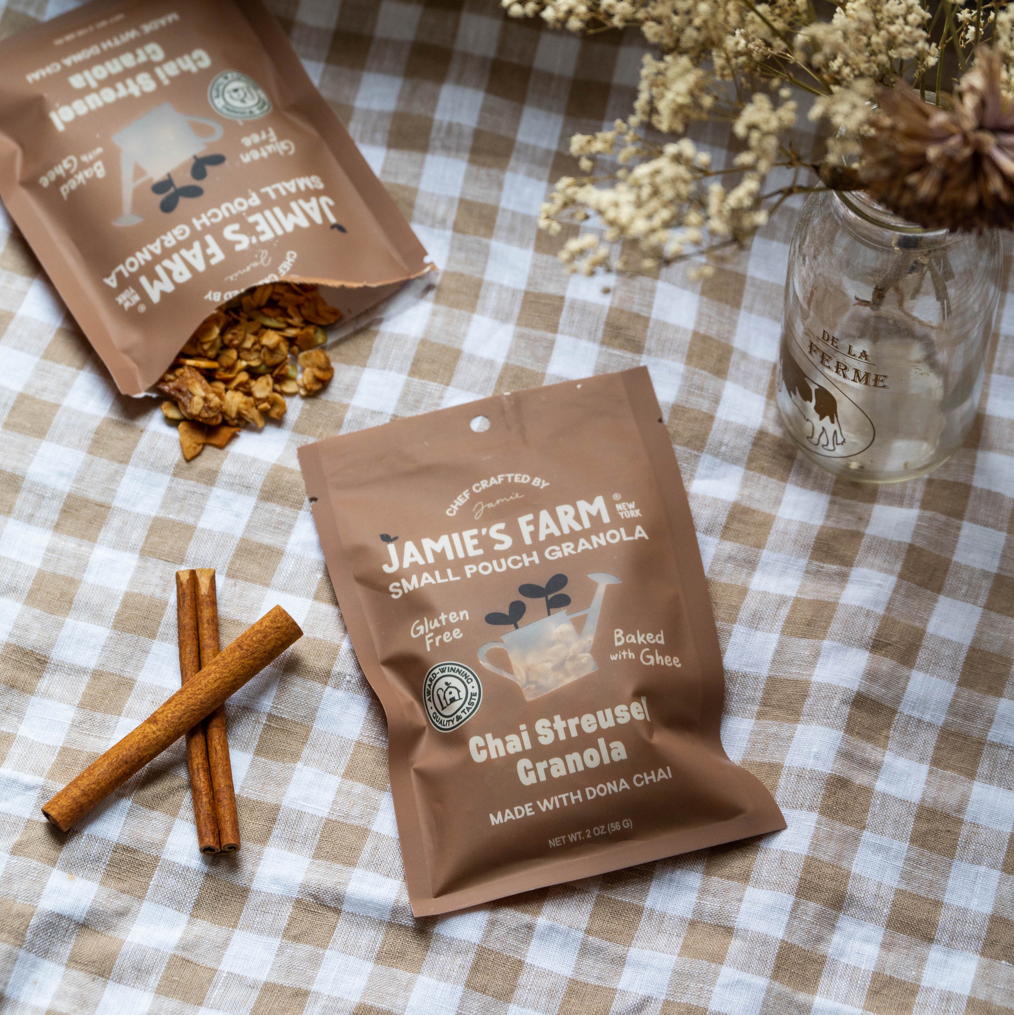 Two bags of Jamie's Farm Small Pouch Granola, specifically Chai Streusel Granola, displayed on a brown and white checkered tablecloth with cinnamon sticks and a glass jar of dried flowers.