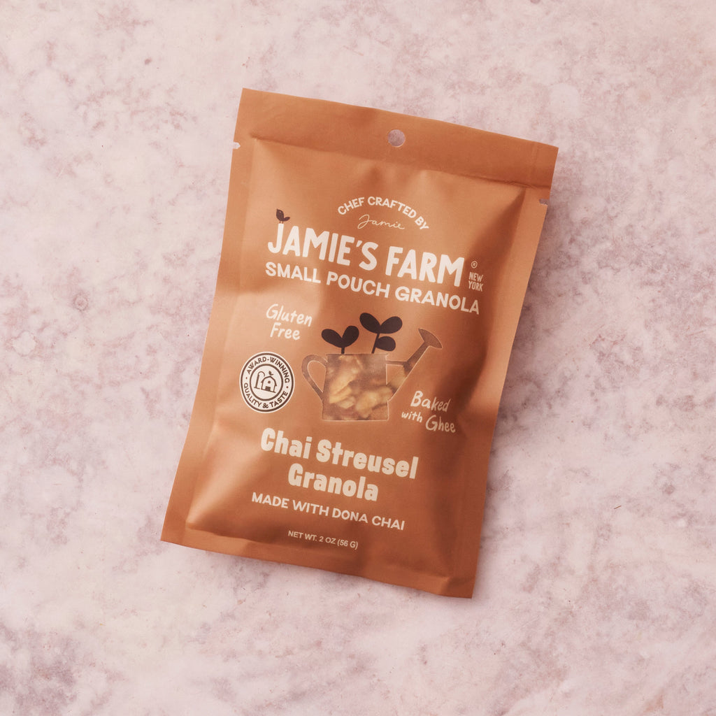 A single pouch of Jamie's Farm Chai Streusel Granola lies on a light pink marble-like surface. The pouch is metallic copper-brown with white text and graphics, including a small illustration of a teacup with leaves.