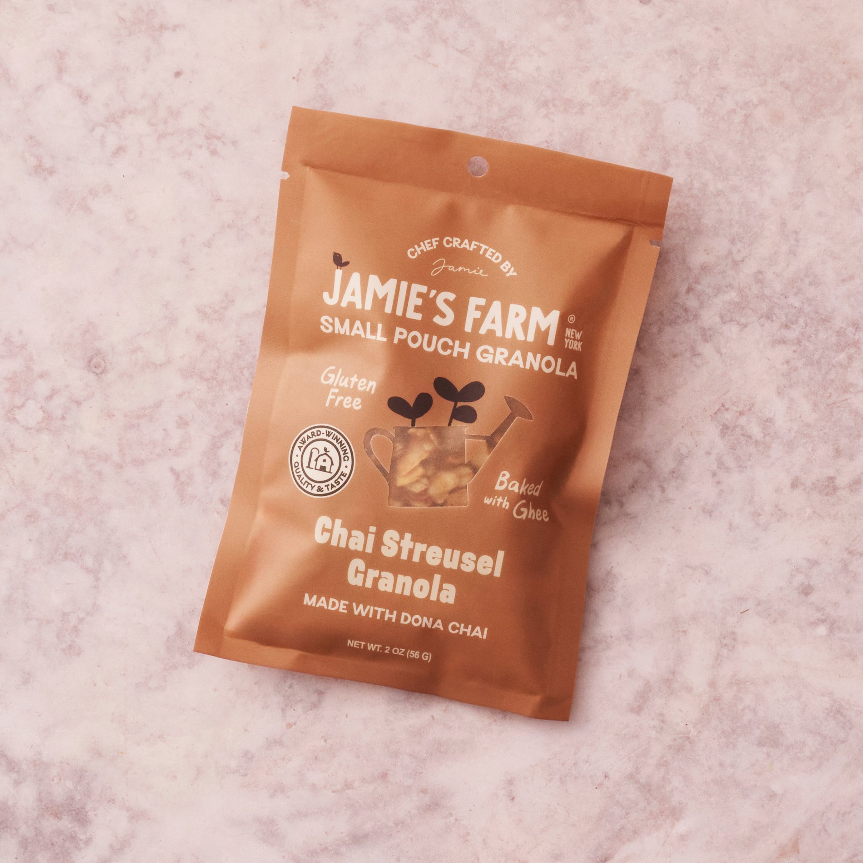 A single pouch of Jamie's Farm Chai Streusel Granola lies on a light pink marble-like surface. The pouch is metallic copper-brown with white text and graphics, including a small illustration of a teacup with leaves.