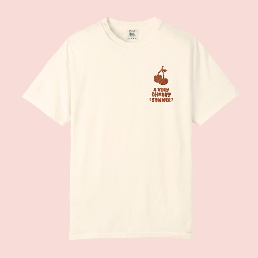 An off-white short-sleeve crewneck t-shirt with a small dark red graphic on the left chest depicting a sailboat and the text 'A VERY CHERRY SUMMER!', displayed against a solid pale pink background.